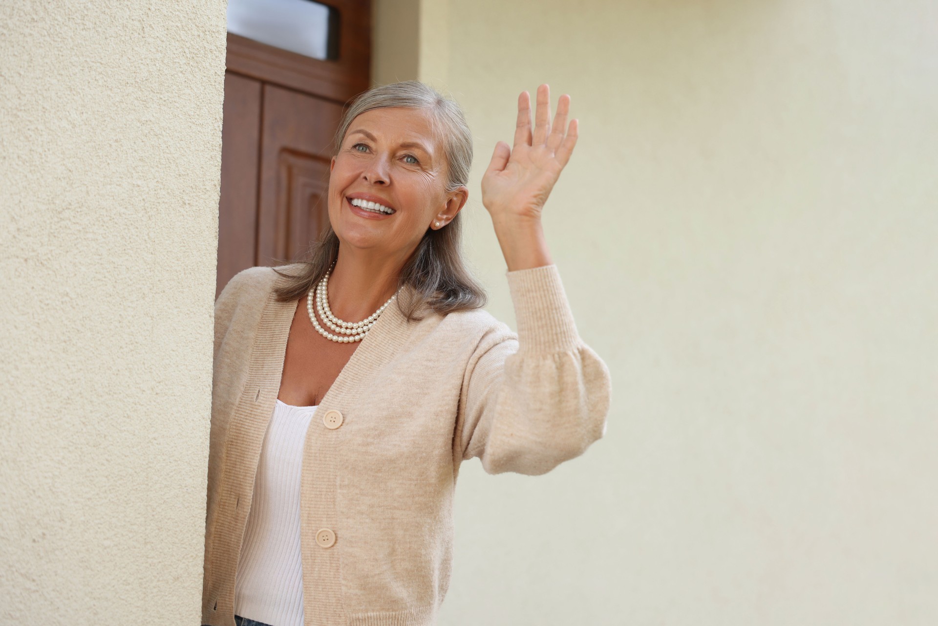 Neighbor greeting. Happy senior woman waving near house outdoors, space for text