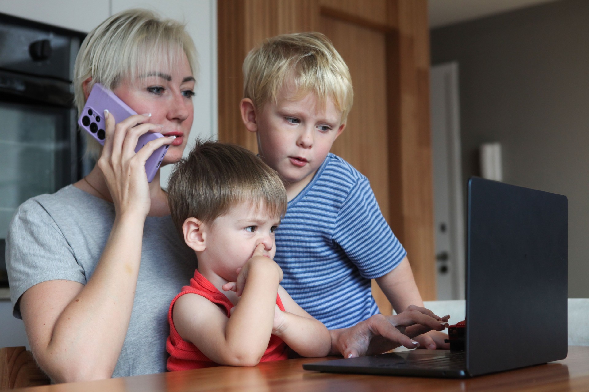 mother talking on cell phone and looking at laptop screen at home with two little  boys